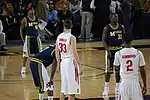 Two opposing basketball teams on a basketball court with a team in dark blue uniforms at the free throw line. One team is in white uniforms with names on the back and the other is dark blue with the word Michigan on the front and names on the back.