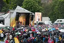  A crowd under umbrellas watches a man on a stage light a cauldron from a torch