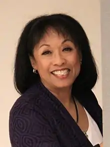 Full body portrait of a women in her late sixties smiling at the camera and with both her hands on a wall