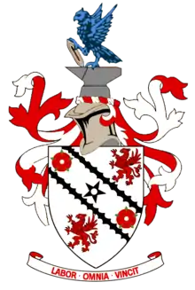 A white shield upon which emblems of two red roses and two red griffins appear at alternate corners. A black five-pointed star is in the centre of the shield's design between two black diagonal lines. Around the shield are red and white ribbons in a symmetrical design. Above the shield is a silver-coloured knight's helmet surmounted by grey anvil upon which a vivid azure-coloured eagle is perched, holding a grey shuttle. Below the shield is the motto "LABOR OMNIA VINCIT".