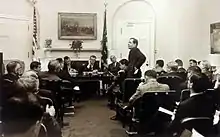 Dr. William Conrad Gibbons (standing), Professional Staff Member of the Democratic Policy Committee and Assistant to the Majority Leader of the Senate, with Senator Lyndon B. Johnson seated in the background