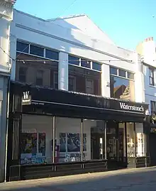 A wide, flat-roofed shop with a pediment. The lower storey has a black shopfront with plate glass windows; the upper storey has three wide rectangular windows. All exposed areas of wall are painted white.