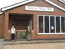 Frontage of a brick-built hall with two young Fulstowians standing by the entrance and the village's war memorial.