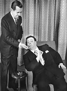 Edward R. Murrow (standing) lighting Gleason's cigarette; Gleason is seated in an armchair