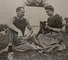 Black and white photo of a middle-aged man and woman wearing casual clothes, sitting outside of a tent, playing with a small dog