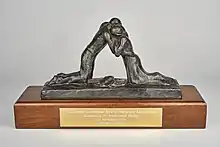 Photograph of The Coventry International Prize for Peace and Reconciliation awarded to Dr Madeleine Sharp in 2004. The award is a metal sculpture of two figures embracing, mounted onto a wooden block with a gold-coloured engraved plaque on the front bearing the inscription "The Coventry International Prize for Peace and Reconciliation Awarded to Dr Madeleine Sharp 14th November 2004 Father Forgive".