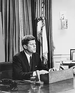 A black and white photograph of President John F. Kennedy speaking into a microphone at his desk in the Oval Office of the White House