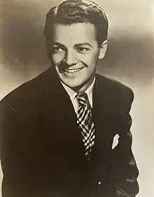 A man wearing a suit and tie is smiling while looking towards the right.