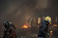 Three helmeted protestors throwing pavement bricks at riot-police line under concealment of smoke from burning tires