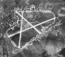 Aerial photograph of the airfield, 2 March 1944. The technical site with two T2 hangars are at the left (west). There are three more T2 hangars and the bomb dump north of the perimeter track. A large number of aircraft are parked on the north/south crosswind runway and on the hardstand loops around the perimeter track