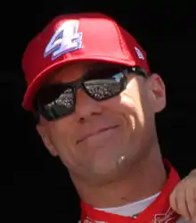 A man in his early forties smiling and waving to the crowd. He is wearing sunglasses along with a red baseball car and similarly colored racing overalls.