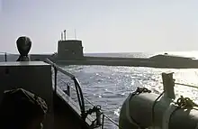 Color photograph depicting the port side of a large surfaced nuclear submarine taken from the bow of an escorting surface ship as the submarine leaves port.