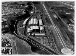Black-and-white aerial photo, facing west toward Pacific Ocean, long runway and hangars surrounded by parked cars, beyond airport are bluffs, roads, fields and homes